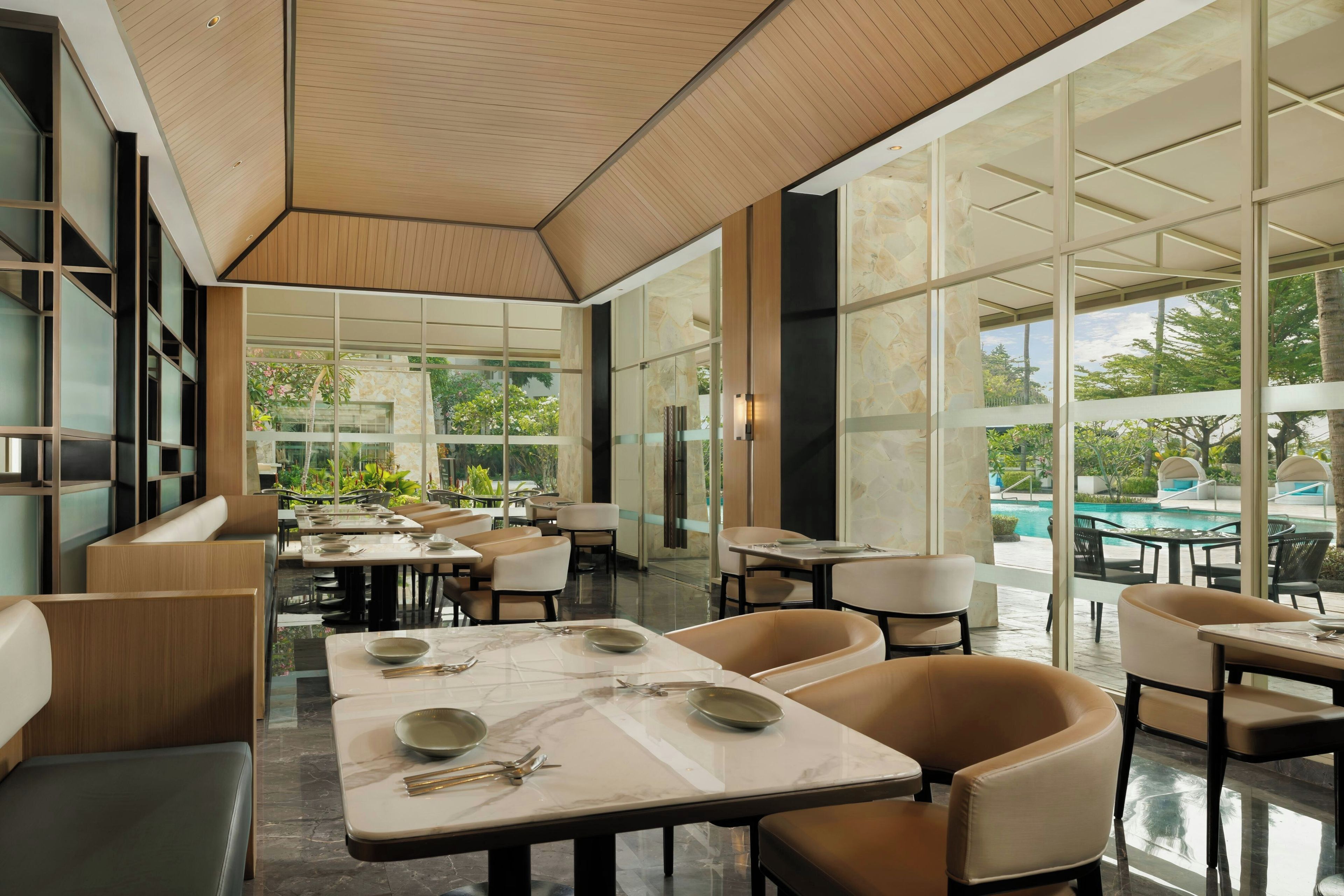 A modern restaurant with glass partitions, marble tables, and a view of a pool and greenery.