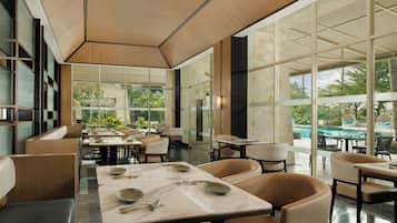 A modern restaurant with glass partitions, marble tables, and a view of a pool and greenery.