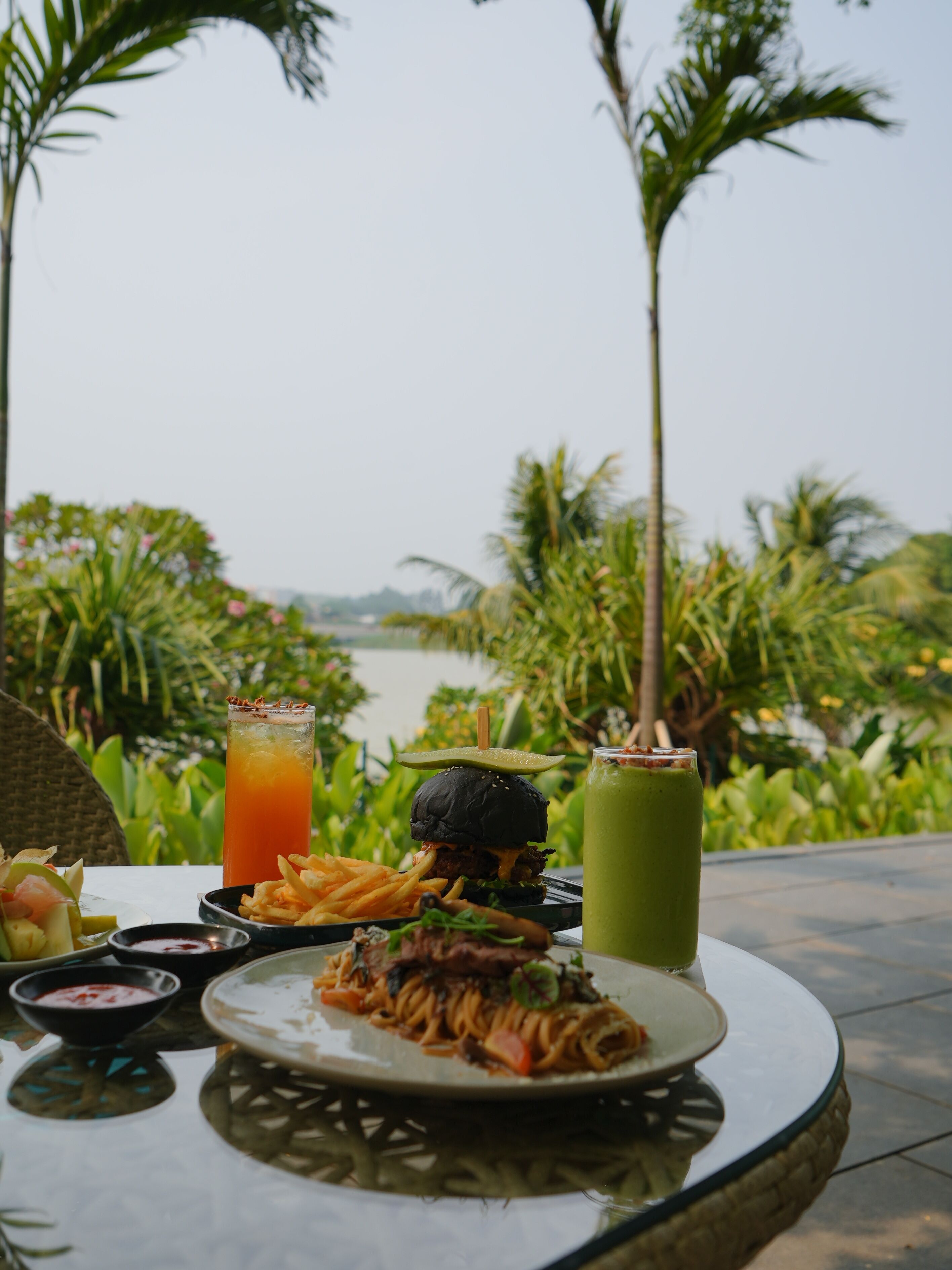 A table with a meal, including a burger, fries, and pasta, and two drinks, set outdoors with a view of a body of water and greenery.