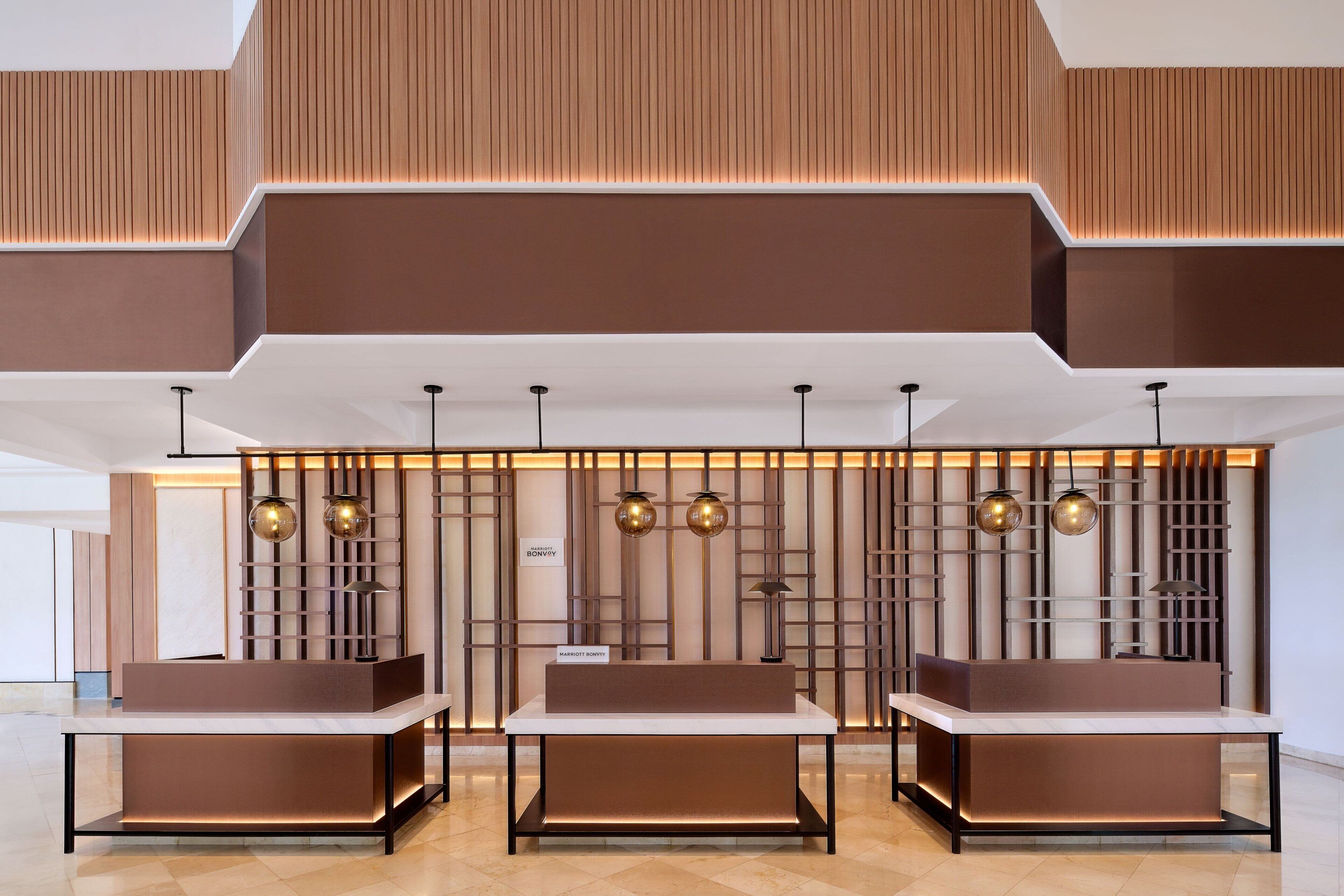Una moderna zona de recepción de hotel con un mostrador central, dos mostradores laterales y una pared decorativa con paneles verticales y luces colgantes.
