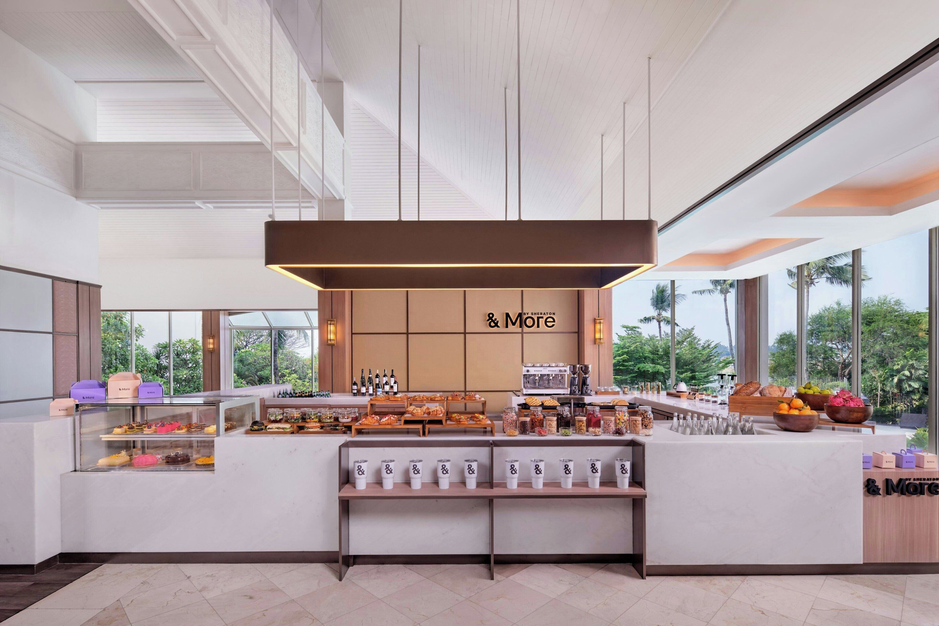 A modern cafe with a central counter displaying pastries and beverages, large windows offering views of greenery, and a minimalist interior design.