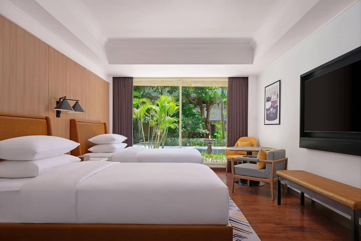 Une chambre d’hôtel avec un grand lit, un bureau, une chaise, un banc, une télévision et une fenêtre donnant sur un paysage verdoyant.