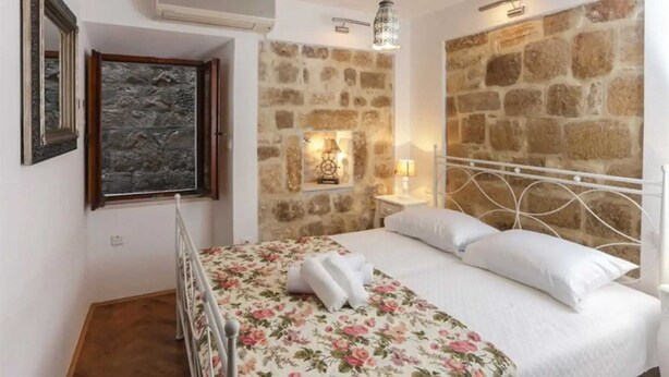 A bedroom with a stone wall, a bed with floral bedding, a nightstand with a lamp, and a hanging light fixture. - Figura suites 2 Studio in the Old town with terrace (Dubrovnik)