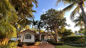 A house with a brick pathway leading to the entrance, surrounded by lush greenery and palm trees.