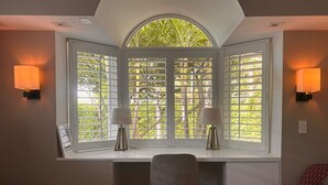 A room with a desk, two wall-mounted lamps, and a window with a view of greenery.