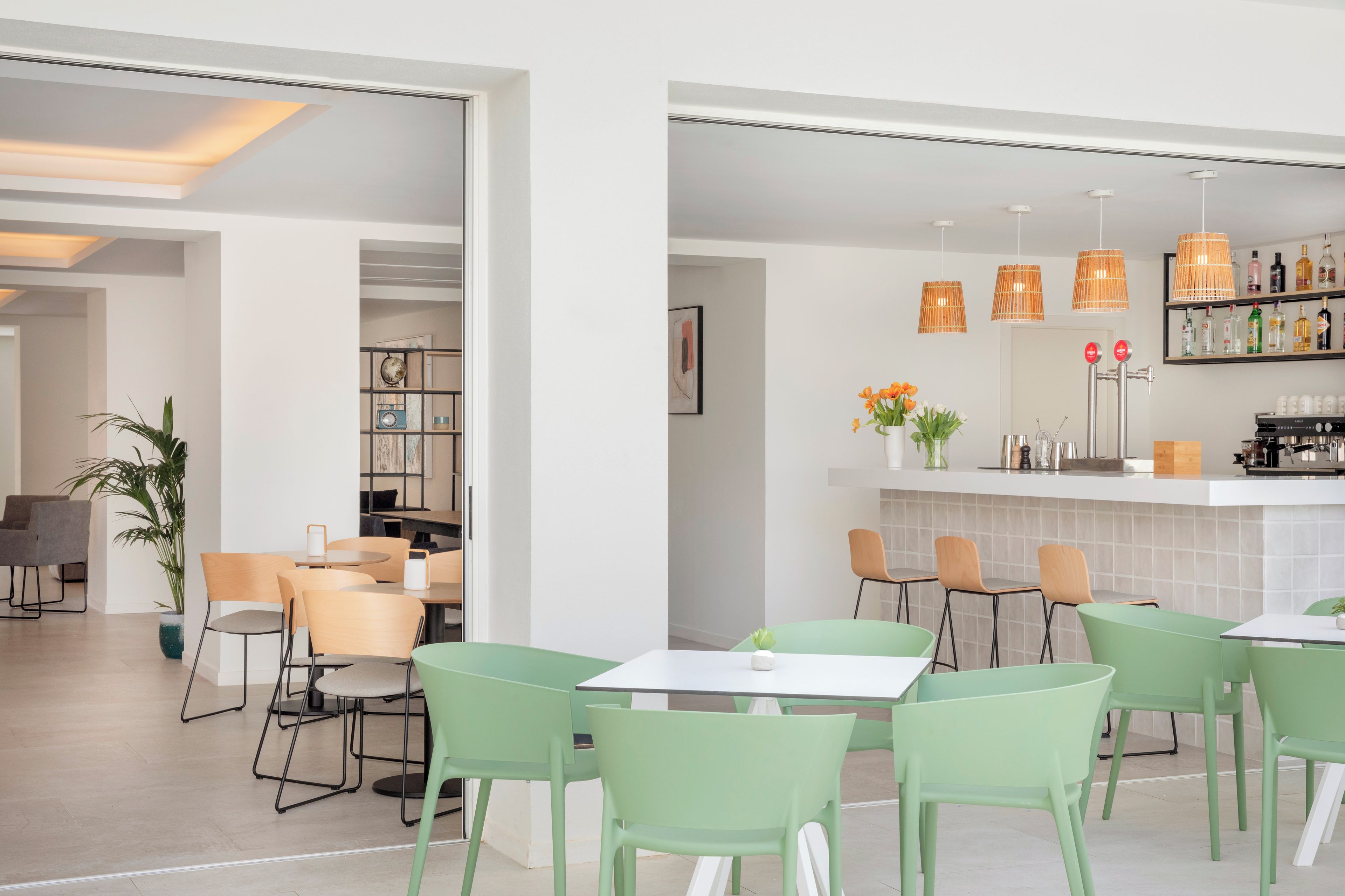 Un espace repas moderne avec des chaises vertes, une table blanche et un coin bar agrémenté de luminaires suspendus et d’étagères remplies de bouteilles.
