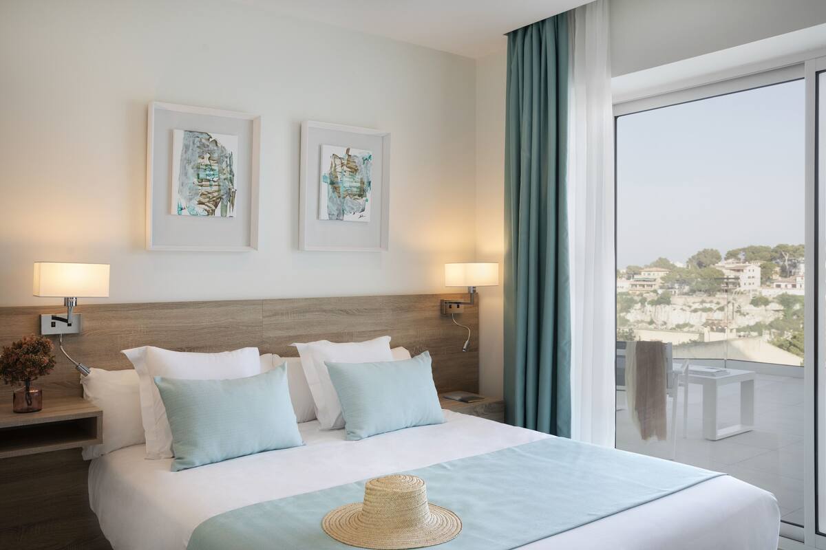 A hotel room with a large bed, two framed artworks on the wall, a balcony with a view, and a hat on the bed.