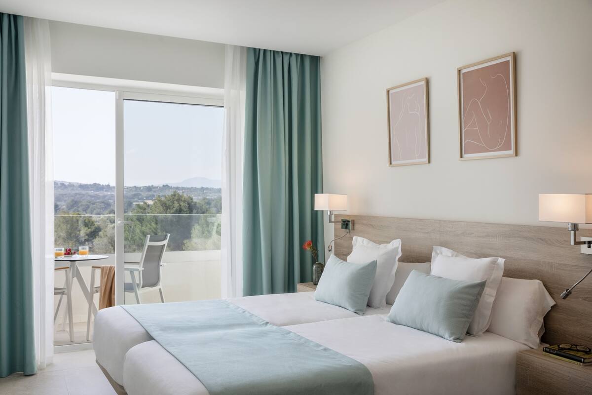 A hotel room with a large bed, two bedside lamps, a view of the outdoors, and a small table with chairs.