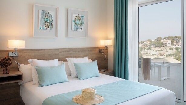A hotel room with a large bed, two framed artworks on the wall, a balcony with a view, and a hat on the bed. - Marina Drach (Manacor)
