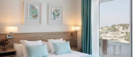 Habitación de hotel con una cama grande, dos cuadros enmarcados en la pared, un balcón con vistas y un sombrero sobre la cama.