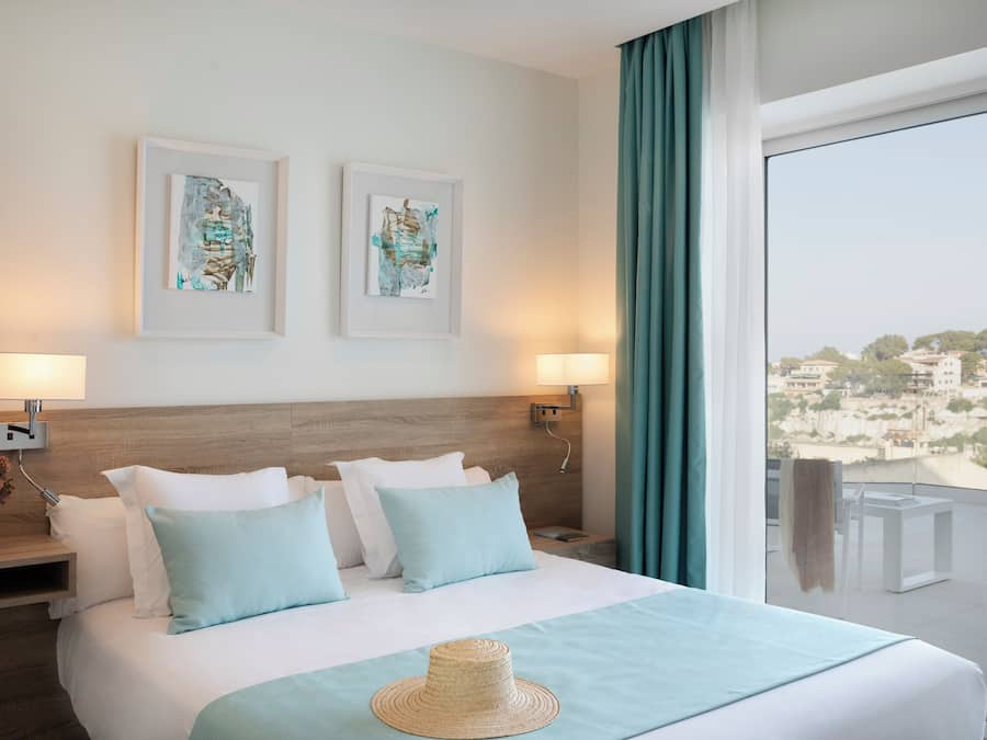 Habitación de hotel con una cama grande, dos cuadros enmarcados en la pared, un balcón con vistas y un sombrero sobre la cama.