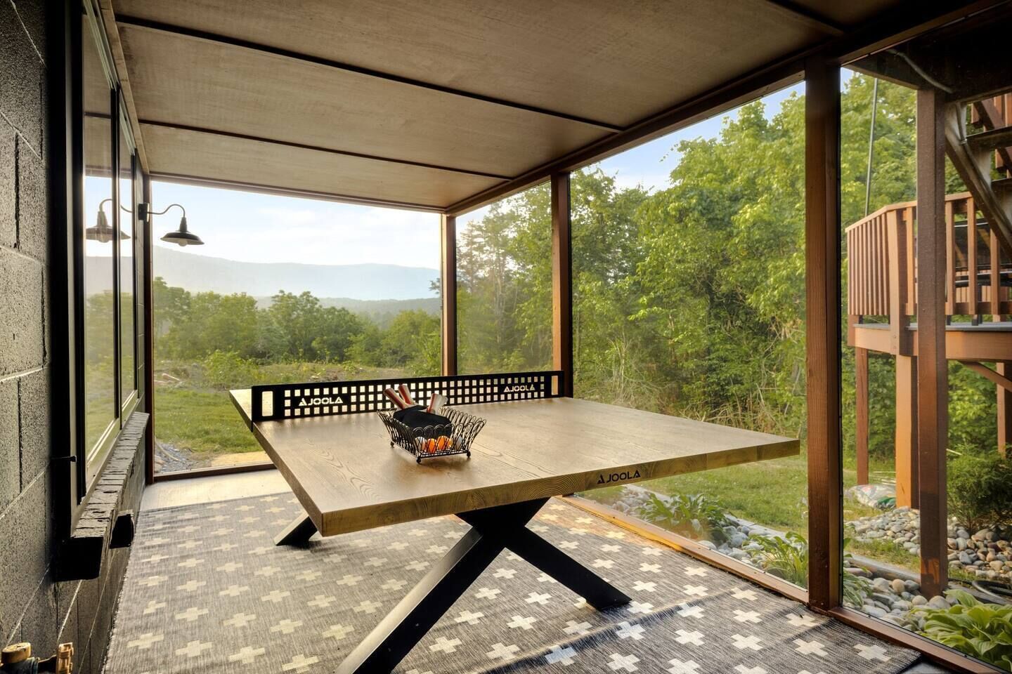 Una mesa de madera con estructura metálica negra, una cesta tejida con naranjas y vistas a un área boscosa a través de amplios ventanales.