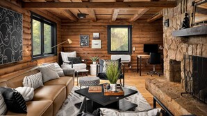 A cozy cabin interior with a wooden ceiling, a stone fireplace, a sofa, a dining table, and a large window. - Luxe Log Cabin, Mountain Views, HotTub, Secluded (Rileyville)
