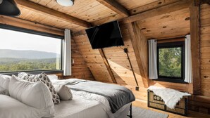 A cozy bedroom with a large bed, a flat-screen TV mounted on the wall, and a view of the outdoors. - Luxe Log Cabin, Mountain Views, HotTub, Secluded (Rileyville)