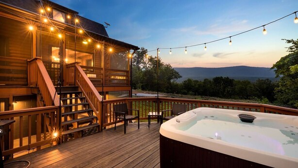 A wooden deck with a hot tub, string lights, and a staircase leading to a covered area. - Luxe Log Cabin, Mountain Views, HotTub, Secluded (Rileyville)
