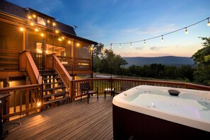 A wooden deck with a hot tub, string lights, and a staircase leading to a covered area. - Luxe Log Cabin, Mountain Views, HotTub, Secluded (Rileyville)