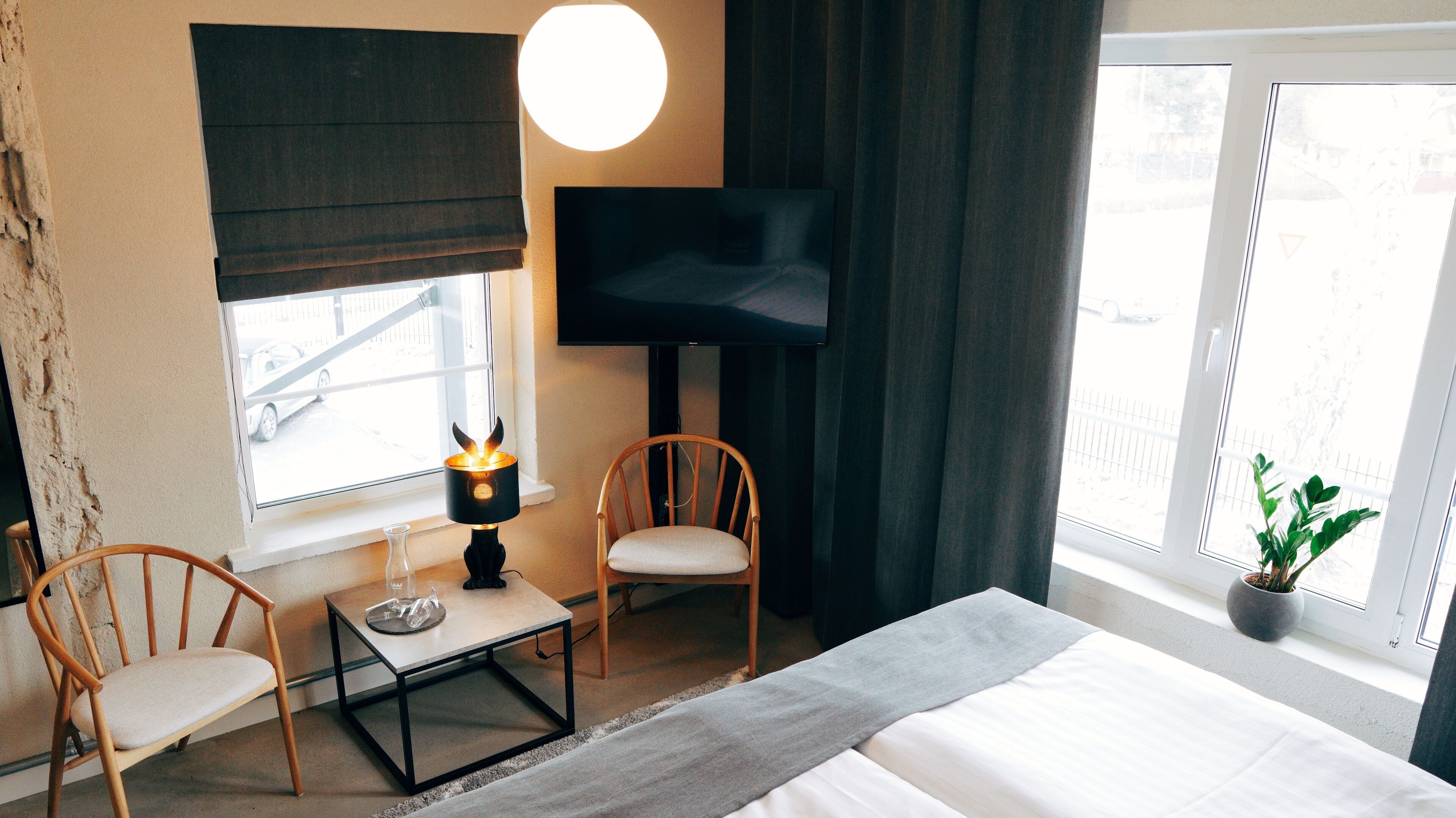 A hotel room with a bed, a TV, a lamp, two chairs, a small table, and a window with a view of the outside.
