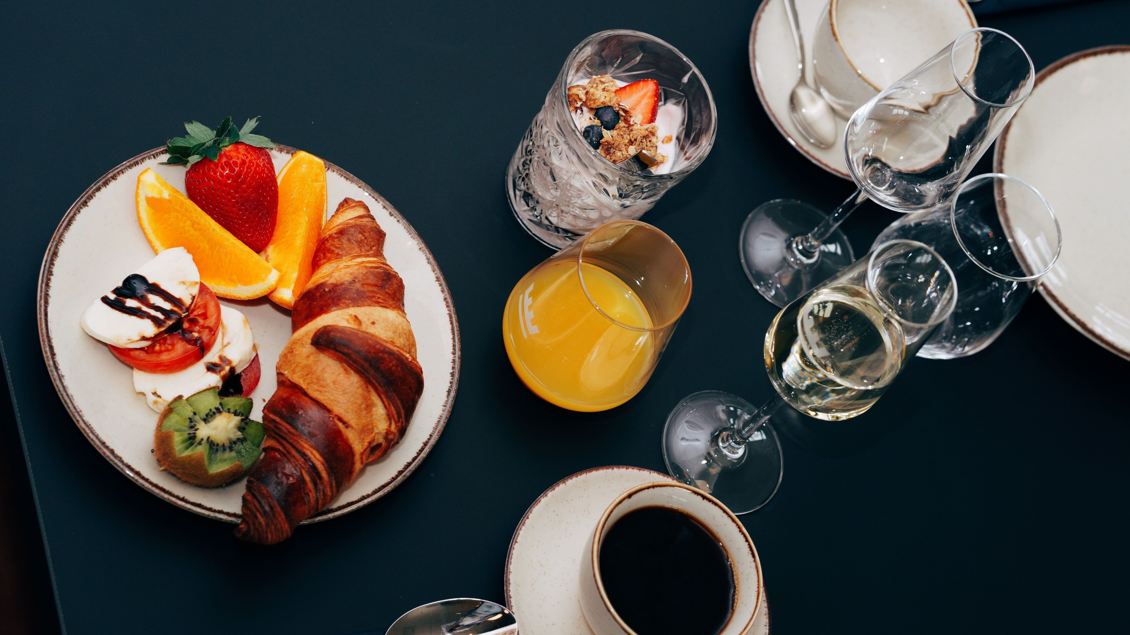 A breakfast setting with a croissant, strawberries, kiwi, orange, and yogurt parfait on a plate, along with glasses of orange juice and white wine.