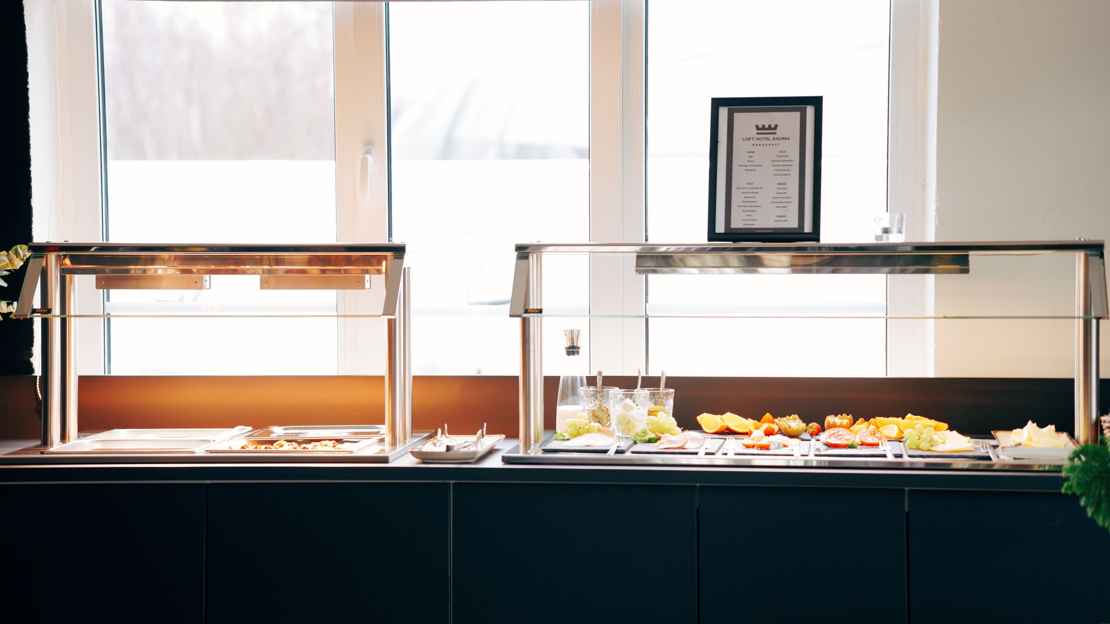 Een ontbijtbuffet met diverse soorten fruit en gebak, een menubord boven de balie en een raam met luxaflex op de achtergrond.