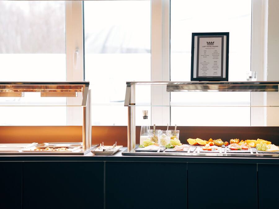 A breakfast buffet with various fruits and pastries, a menu board above the counter, and a window with blinds in the background.