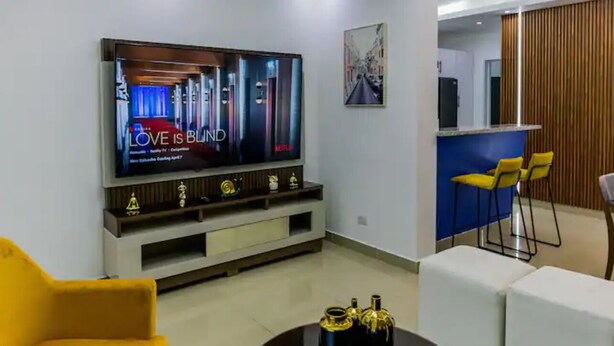 A modern living room with a large flat-screen TV, a yellow armchair, a white sofa, and a small round table with decorative bottles. - Helios Luxurious Condo At Caleta (La Romana)