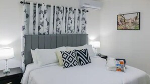 A hotel room with a bed, white bedding, a grey headboard, patterned curtains, a wall-mounted air conditioner, a bedside table with a lamp, and a framed picture on the wall. - Helios Luxurious Condo At Caleta (La Romana)