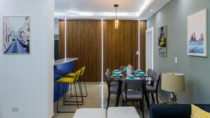 A modern living room with a dining area, featuring a sofa, a dining table with chairs, and a lamp. - Helios Luxurious Condo At Caleta (La Romana)