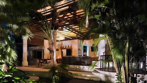 Un espacio exterior bien iluminado con piscina, palmeras y una zona de estar cubierta.