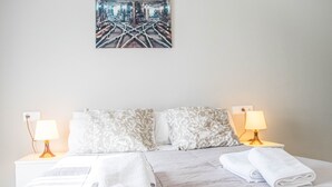 Una cama bien hecha con sábanas y almohadas blancas, dos lámparas de noche y una obra enmarcada sobre la cama.