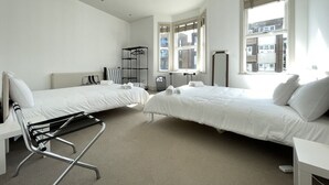 Une chambre d’hôtel avec deux lits, un radiateur, une chaise et une fenêtre donnant sur des immeubles.