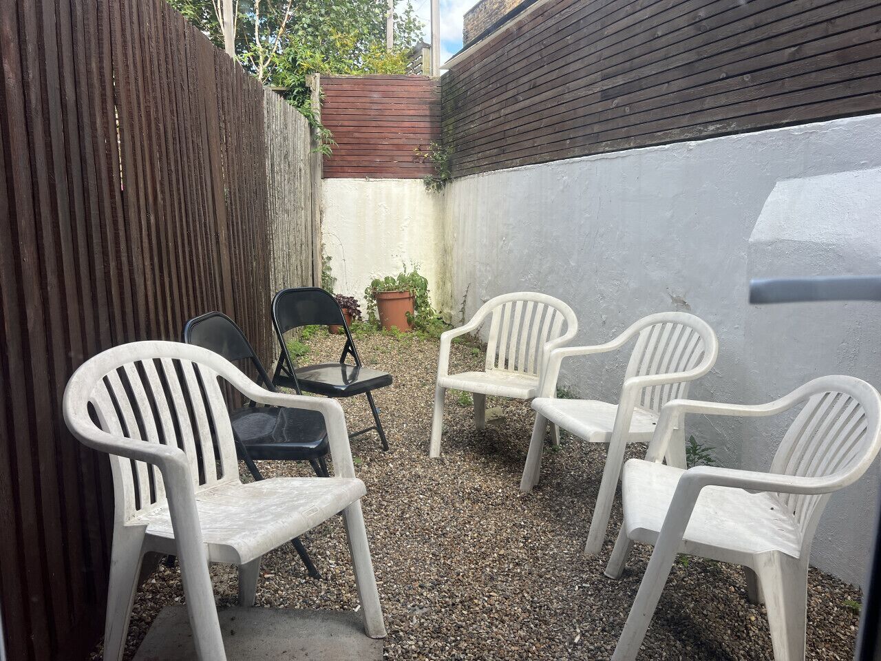 A small outdoor seating area with four white plastic chairs and one black chair, surrounded by a gravel ground and a wooden fence.