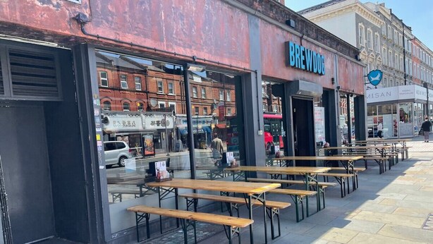 Un espace extérieur aménagé avec des tables et des bancs en bois, une devanture de magasin portant l’enseigne « BREWDOG », et un ciel dégagé en arrière-plan.