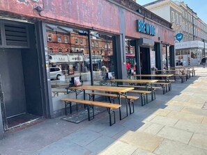 An outdoor seating area with wooden tables and benches, a storefront with a sign reading "BREWDOG," and a clear sky in the background. - Sleeps 16 - 4BR London Home - Near Westfield & Metro (Greater London)