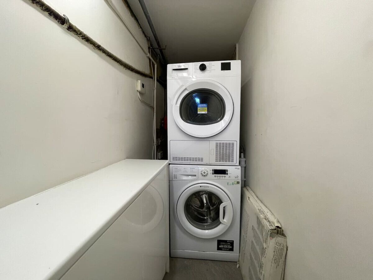 Ein kleiner Hauswirtschaftsraum mit einer übereinander angeordneten Waschmaschine und einem Trockner, einer weißen Wand und einem sichtbaren Rohr.