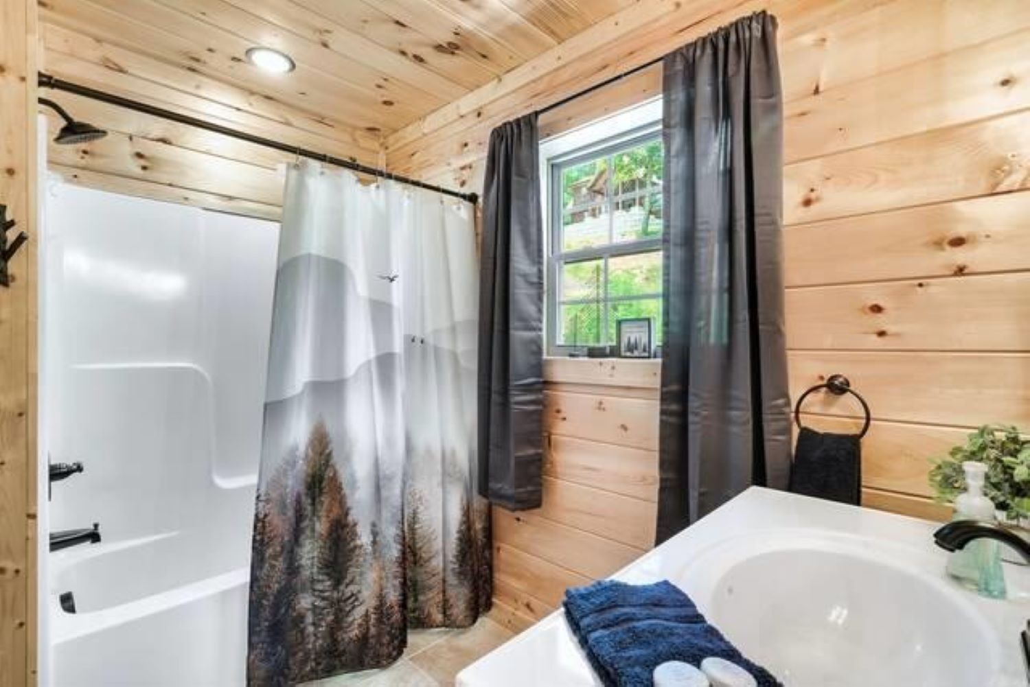 A wooden cabin bathroom with a bathtub, a shower curtain with a forest design, and a window with a view of greenery.