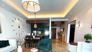 Una sala de estar moderna con una mesa de centro de madera, un sillón color turquesa y un sofá blanco. Se ve un comedor al fondo con una cocina.