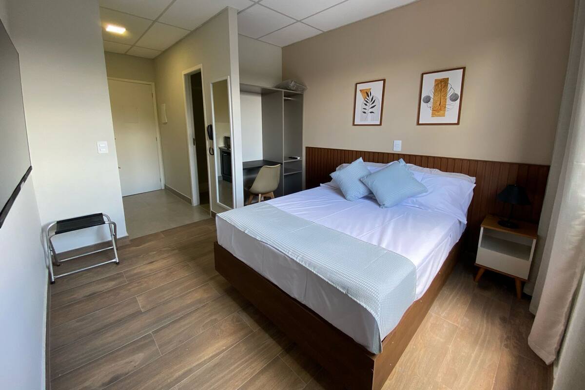 A hotel room with a large bed, a desk, and a chair. There is a wardrobe and a small table with a lamp.