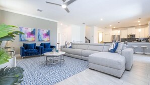 Un salon moderne avec un canapé d’angle, une table basse et un tapis à motifs. On aperçoit à l’arrière-plan un coin cuisine avec un réfrigérateur et un four à micro-ondes.