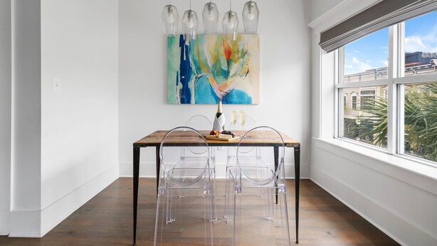 A dining area with a wooden table, clear chairs, a colorful abstract painting, and a view through a large window. - The Heart of Historic King St | Cooper Suite (Charleston)