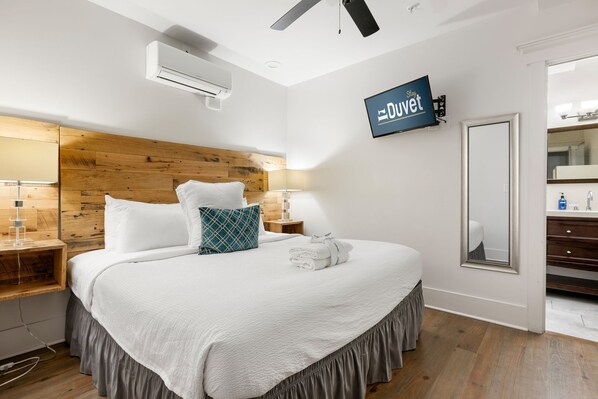 A hotel room with a large bed, wooden headboard, white bedding, a flat-screen TV mounted on the wall, a mirror, and a bathroom with a sink and mirror.