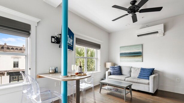 Een moderne woonkamer met een witte bank, een glazen eettafel en een transparante stoel. Er is een plafondventilator, een raam met uitzicht op gebouwen en een airconditioner aan de muur.