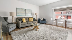 A modern living room with a grey sofa, two armchairs, a glass coffee table, and a large window with a view of a building. - In the Heart of King Street | Hampton Suite (Charleston)