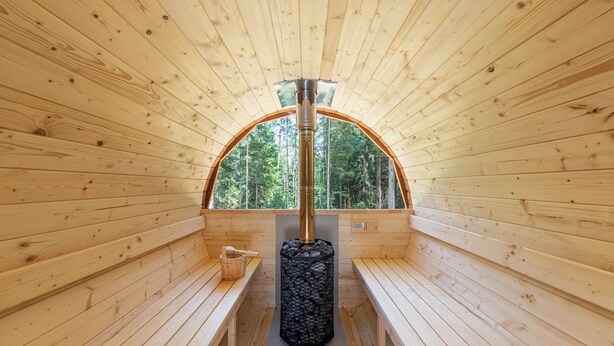 A sauna with wooden walls and benches, a central stone pillar, and a view of trees through an arched window. - C_AL RANCH WELLNESS CORTINA, LAKE CADORE, DOLOMITES (PIEVE DI CADORE)