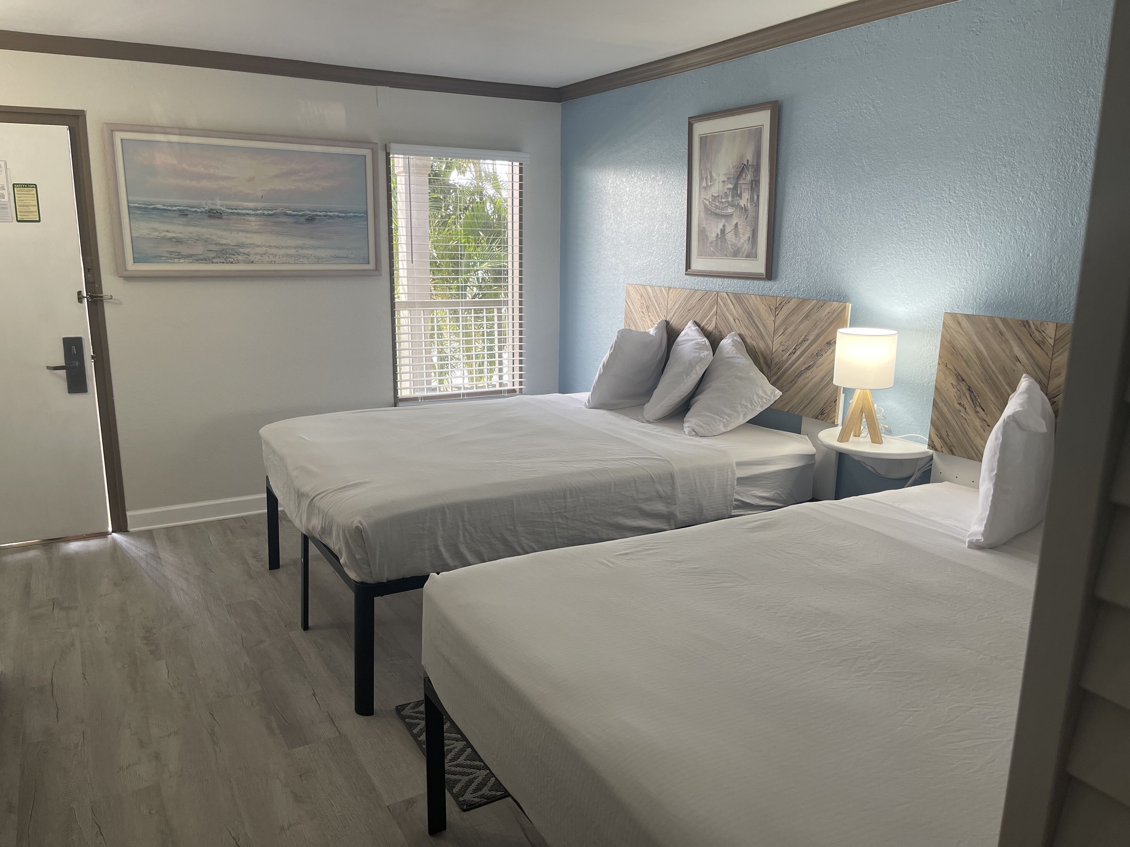 A hotel room with two beds, a wooden headboard, a nightstand with a lamp, and two framed pictures on the wall.