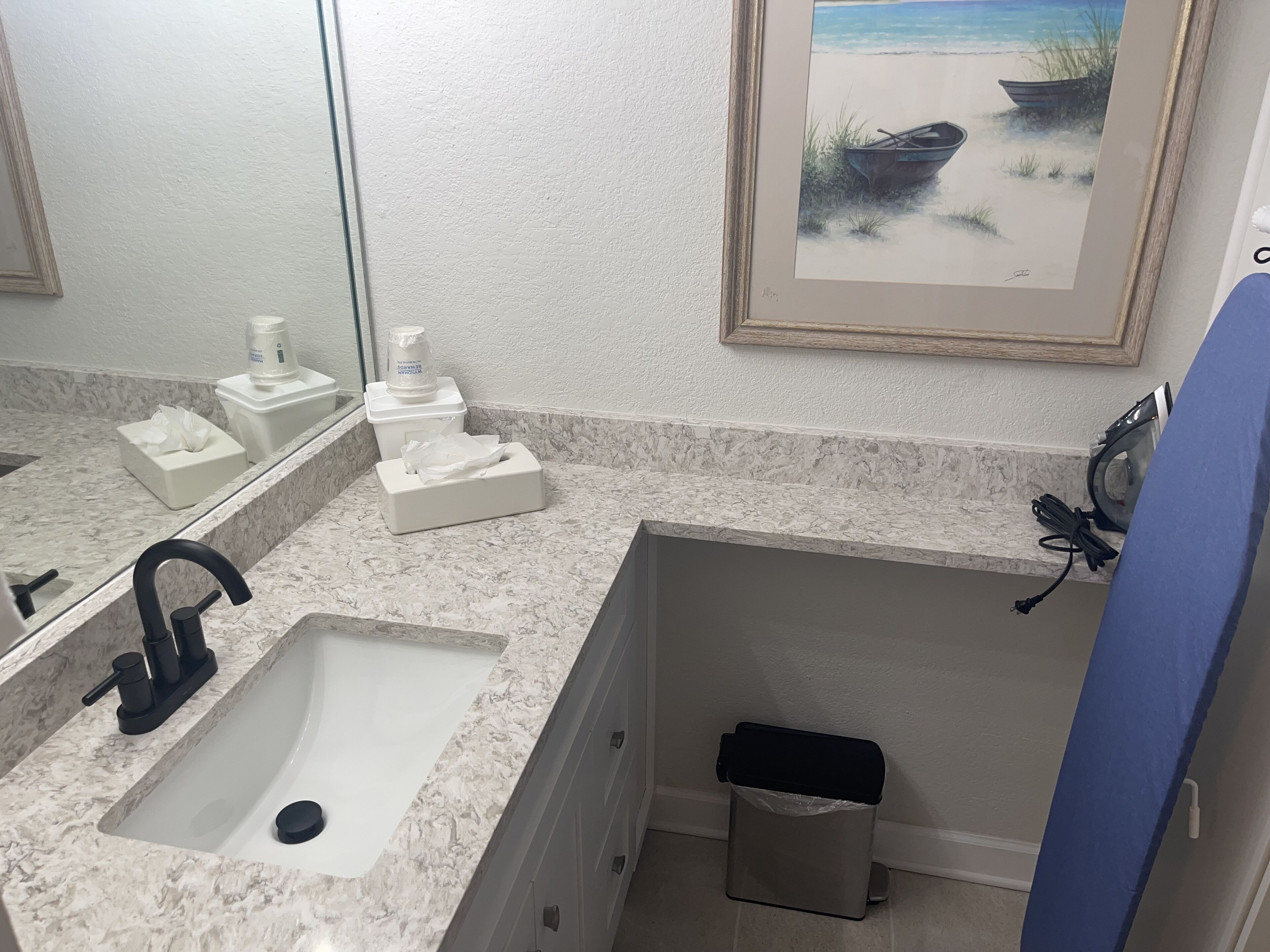 A bathroom with a marble sink, a mirror, a framed beach painting, and a trash bin.