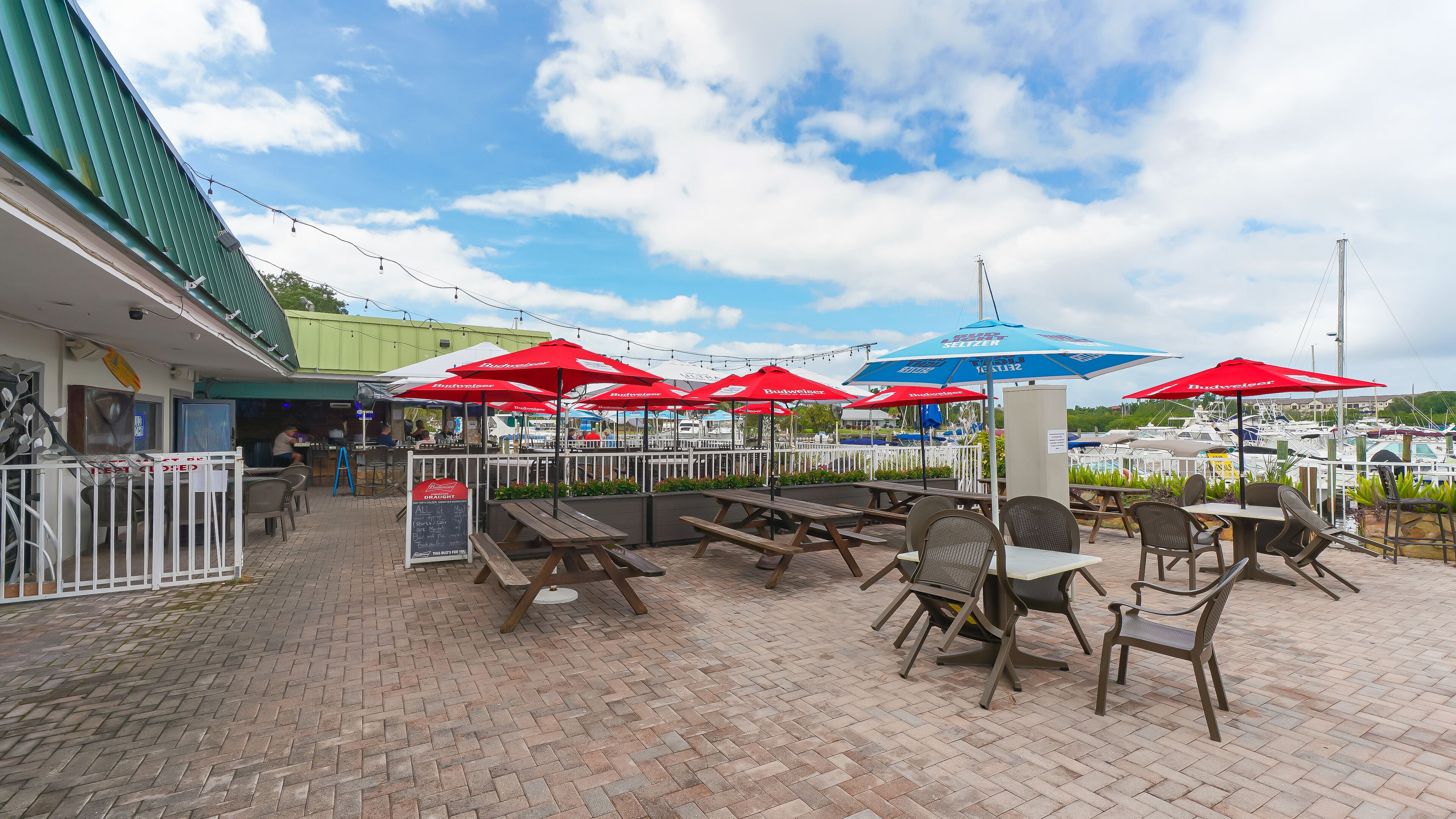 An outdoor seating area with tables and chairs, red and blue umbrellas, and a view of boats in the background.