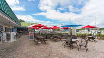 An outdoor seating area with tables and chairs, red and blue umbrellas, and a view of boats in the background.