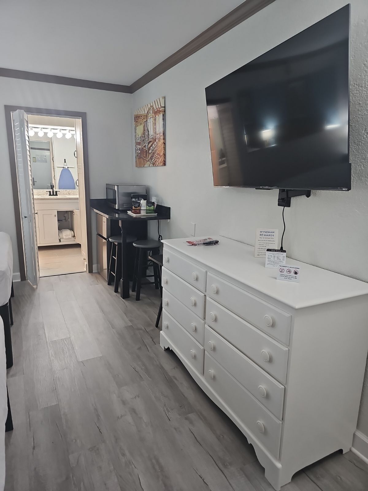 Hotelkamer met een grote flatscreen televisie aan de muur, een witte dressoir met lades, een bureau met een computer en stoel, en een badkamer met spiegel en wastafel.