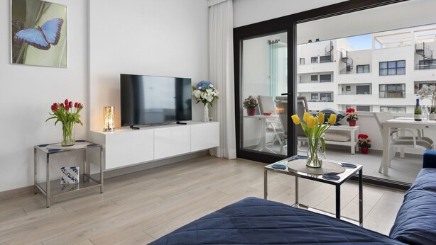 Una sala de estar moderna con un televisor de pantalla plana, un sofá, una mesa de centro con un jarrón de flores y un balcón con vista a la ciudad.