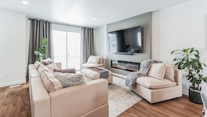 Una sala de estar moderna con un sofá beige, una mesa de centro y un televisor de pantalla plana montado en la pared. Hay un ventanal con cortinas, una planta en maceta y una chimenea.